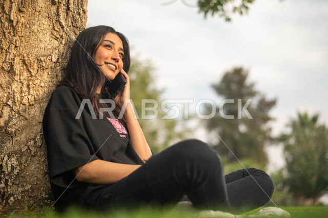 إستخدام الهاتف المحمول, صورة جانبية لشابة عربية خليجية سعودية تجلس تحت شجرة في حديقة عامة و تقوم بالرد على الاتصالات الهاتفية, إيماءات وجه تدل على السعادة، نزهه في الطبيعة الخضراء، قضاء أوقات ممتعة في الهواء الطلق.