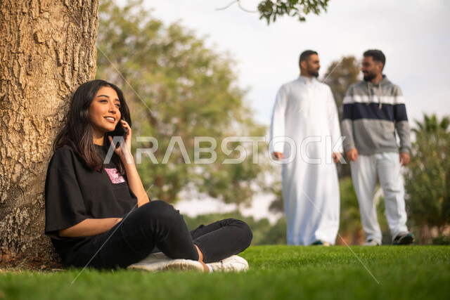 شابة عربية خليجية سعودية تجلس تحت الشجرة و تقوم بإجراء اتصال هاتفي, إيماءات وجه تدل على السعادة، نزهه في الطبيعة الخضراء وقت النهار، الرد على المكالمات الهاتفية, شابان عربيان خليجيان سعوديان يتجولان في الحديقة العامة, قضاء أوقات ممتعة في الهواء الطلق.