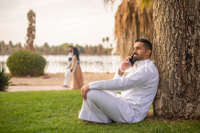 رجل عربي خليجي سعودي يجلس تحت شجرة و يقوم بإجراء الاتصالات الهاتفية, استخدام الهاتف المحمول،الحدائق العامة في المملكة العربية السعودية، إيماءات وجه تدل على السعادة، نزهه في الهواء الطلق، قضاء أوقات ممتعة في الطبيعة الخضراء.