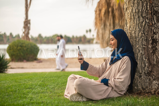 إمرأة عربية خليجية سعودية تجلس تحت شجرة و تجري مكالمة فيديو عبر الهاتف المحمول، الحدائق العامة في المملكة العربية السعودية، إيماءات وجه تدل على السعادة، نزهه في الطبيعة الخضراء ، قضاء أوقات ممتعة في الهواء الطلق.