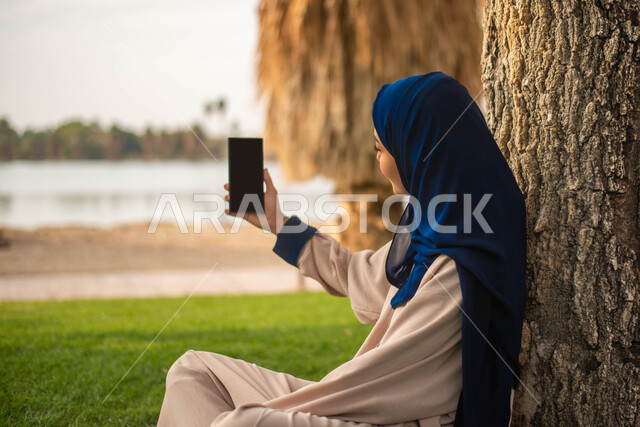 نزهه في الهواء الطلق و الطبيعة الخضراء, التقاط صور تذكارية باستخدام الهاتف المحمول، صورة جانبية لإمرأة عربية خليجية سعودية تجلس على العشب و تقضي أوقات ممتعة في إحدى حدائق المملكة العربية السعودية، بث مباشر.