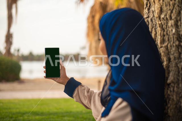 نزهه في الهواء الطلق و الطبيعة الخضراء, التقاط صور تذكارية باستخدام الهاتف المحمول، صورة جانبية لإمرأة عربية خليجية سعودية تقضي أوقات ممتعة في إحدى حدائق المملكة العربية السعودية،  بث مباشر.