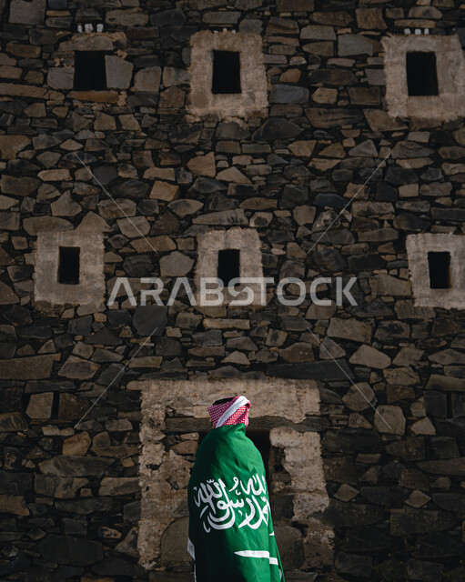 صورة من الخلف لرجل عربي سعودي خليجي يرتدي علم السعودية احتفالا بذكرى اليوم الوطني السعودي في قرية رجال ألمع في عسير في المملكة العربية السعودية، يوم العلم السعودي، العيد الوطني السعودي، يوم التأسيس السعودي، بيوت ومباني اثرية تاريخية قديمة
