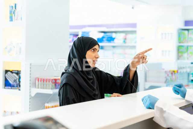 سيدة عربية خليجية سعودية تقوم بطلب الادوية من صيدلي خليجي يرتدي الكمامة للوقاية من فايروس كورونا ، اختيار الادوية والمنتجات الطبية ،شراء الادوية الموجودة في الوصفات الطبية  ، الاستفادة من الخدمات والرعاية الصحية 