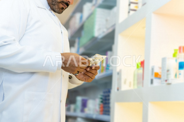 رجل صيدلاني عربي سعودي خليجي يحمل بيده الدواء ، يقوم بجرد الادوية ، فحص مخزون الأدوية ، تقديم الرعاية الصحية بجودة عالية ، الادوية والمستلزمات الطبية ، مهنة الصيدلة