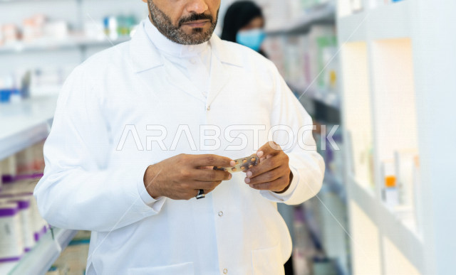 رجل صيدلاني عربي سعودي خليجي يحمل بيده الدواء ، يقوم بجرد الادوية ، فحص مخزون الأدوية ، تقديم الرعاية الصحية بجودة عالية ، الادوية والمستلزمات الطبية ، مهنة الصيدلة