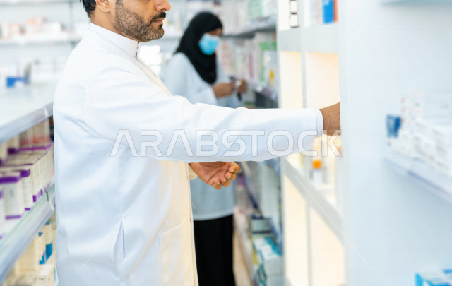 رجل وامراة عربيان خليجيان سعوديان يرتديان الكمامة للوقاية من فايروس كورونا ،  يقومان بجرد الادوية داخل الصيدلية ، مناقشة امور خاصة بالعمل ، فحص مخزون الأدوية ، تقديم الرعاية الصحية بجودة عالية ، الادوية والمستلزمات الطبية ، مهنة الصيدلة