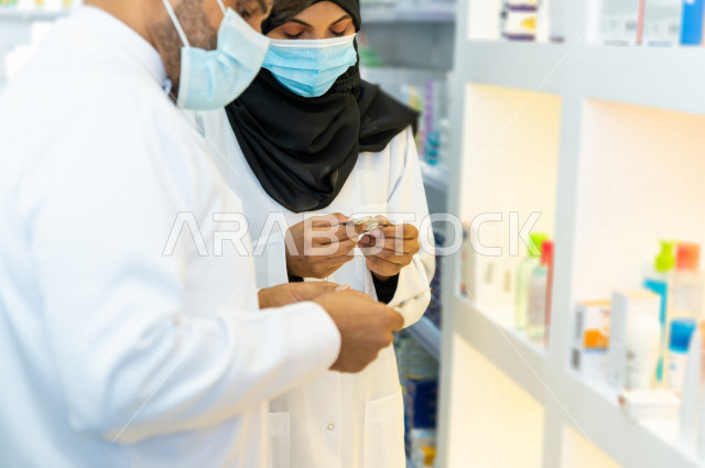 رجل وامراة عربيان خليجيان سعوديان يرتديان الكمامة للوقاية من فايروس كورونا ،  يقومان بجرد الادوية داخل الصيدلية ، مناقشة امور خاصة بالعمل ، فحص مخزون الأدوية ، تقديم الرعاية الصحية بجودة عالية ، الادوية والمستلزمات الطبية ، مهنة الصيدلة