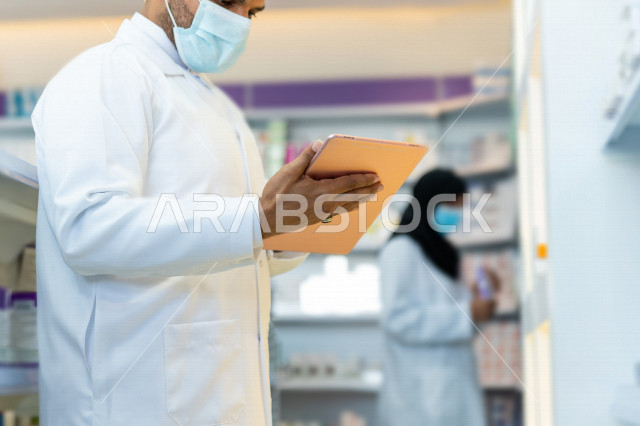 رجل صيدلاني عربي سعودي خليجي يرتدي الكمامة للوقاية من فايروس كورونا ، يقوم بجرد الادوية داخل الصيدلية ، فحص مخزون الأدوية ، استخدام جهاز التابلت لتدوين الملاحظات ، تقديم الرعاية الصحية بجودة عالية ، الادوية والمستلزمات الطبية ، مهنة الصيدلة