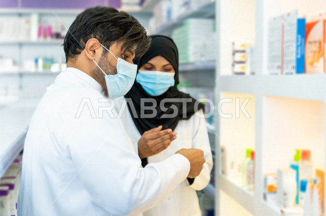 رجل وامراة عربيان خليجيان سعوديان يرتديان الكمامة للوقاية من فايروس كورونا ،  يقومان بجرد الادوية داخل الصيدلية ، مناقشة امور خاصة بالعمل ، فحص مخزون الأدوية ، تقديم الرعاية الصحية بجودة عالية ، الادوية والمستلزمات الطبية ، مهنة الصيدلة