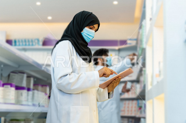 مرأه صيدلانية عربية سعودية خليجية ترتدي الكمامة للوقاية من فايروس كورونا  ، تقوم بجرد الادوية داخل الصيدلية ،  فحص مخزون الأدوية و تدوين الملاحظات على جهاز التابلت ، تقديم الرعاية الصحية بجودة عالية ، الادوية والمستلزمات الطبية ، مهنة الصيدلة 