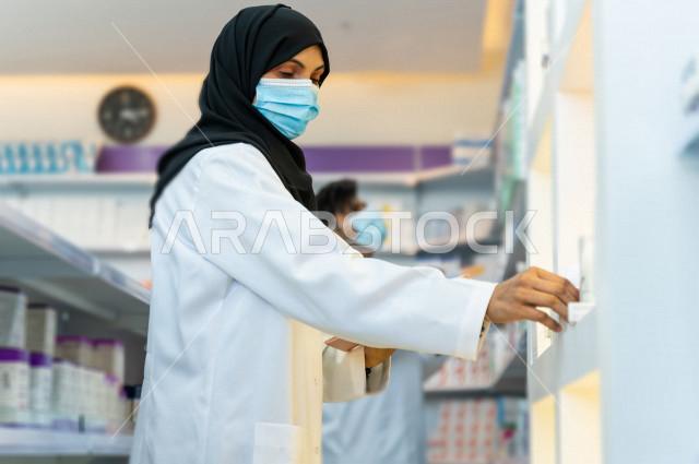 A Saudi Arabian Gulf woman pharmacist wears a mask to prevent corona virus, inventorying medicines inside the pharmacy, checking drug stocks and taking notes on the tablet device, providing high-quality health care, medicines and medical supplies, pharmac