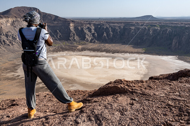 صورة من الخلف لشاب عربي خليجي سعودي يلتقط الصور من خلال الكاميرا لفوهة الوعبة في مدينة الطائف في السعودية، حفرة الوعبة البركانية، اماكن تاريخية أثرية، الحمم البركانية على منحدر فوهة الوعبة، السياحية في السعودية