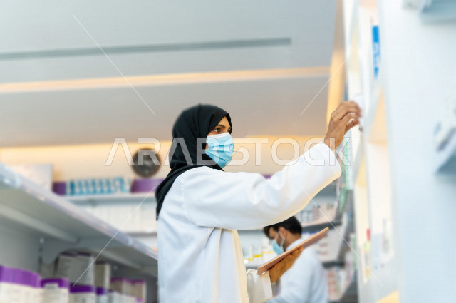 مرأه صيدلانية عربية سعودية خليجية ترتدي الكمامة للوقاية من فايروس كورونا  ، تقوم بجرد الادوية داخل الصيدلية ،  فحص مخزون الأدوية و تدوين الملاحظات على جهاز التابلت ، تقديم الرعاية الصحية بجودة عالية ، الادوية والمستلزمات الطبية ، مهنة الصيدلة 