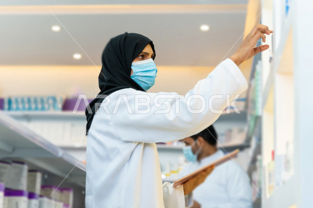 مرأه صيدلانية عربية سعودية خليجية ترتدي الكمامة للوقاية من فايروس كورونا  ، تقوم بجرد الادوية داخل الصيدلية ،  فحص مخزون الأدوية و تدوين الملاحظات على جهاز التابلت ، تقديم الرعاية الصحية بجودة عالية ، الادوية والمستلزمات الطبية ، مهنة الصيدلة 