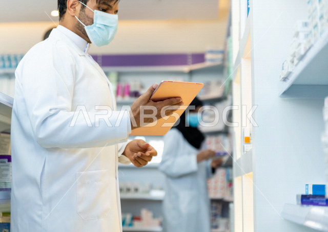 رجل صيدلاني عربي سعودي خليجي يرتدي الكمامة للوقاية من فايروس كورونا ، يقوم بجرد الادوية داخل الصيدلية ، فحص مخزون الأدوية ، استخدام جهاز التابلت لتدوين الملاحظات ، تقديم الرعاية الصحية بجودة عالية ، الادوية والمستلزمات الطبية ، مهنة الصيدلة