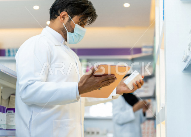 رجل صيدلاني عربي سعودي خليجي يرتدي الكمامة للوقاية من فايروس كورونا ، يقوم بجرد الادوية داخل الصيدلية ، فحص مخزون الأدوية ، استخدام جهاز التابلت لتدوين الملاحظات ، تقديم الرعاية الصحية بجودة عالية ، الادوية والمستلزمات الطبية ، مهنة الصيدلة