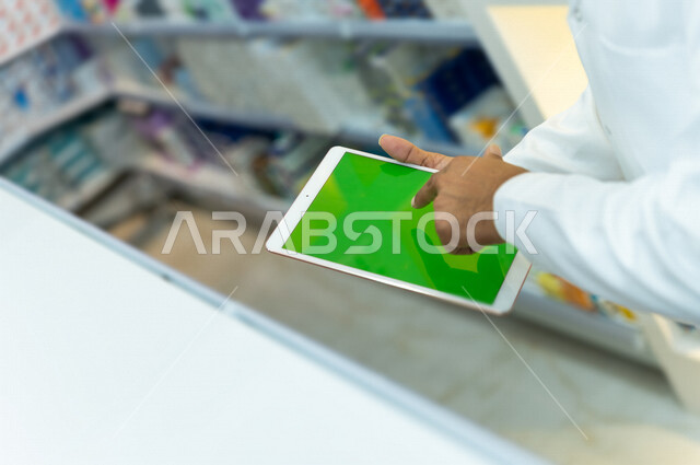 صيدلي عربي خليجي سعودية يحمل بيده جهاز التابلت بشاشة كروما الخضراء  ، فحص مخزون الادوية ، عرض الادوية والمنتجات الطبية ، بيع الادوية اونلاين ، الاعلان والتسويق للمستلزمات الطبية ، مهنة الصيدلة 