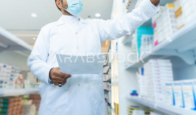 رجل صيدلاني سعودي خليجي يعمل في الصيدلية ، يرتدي الكمامة للوقاية من فايروس كورونا ،  يقوم بفحص مخزون الأدوية ويتحقق من صحتها ، الادوية والمستلزمات الطبية ، مهنة الصيدلة 