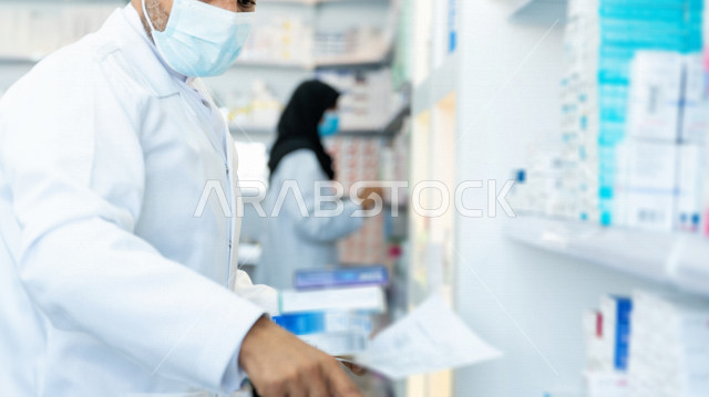 رجل وامراة عربيان خليجيان سعوديان يرتديان الكمامة للوقاية من فايروس كورونا ،  يقومان بجرد الادوية داخل الصيدلية ، تدوين الملاحظات الخاصة بالادوية ، فحص مخزون الأدوية ، تقديم الرعاية الصحية بجودة عالية ، الادوية والمستلزمات الطبية ، مهنة الصيدلة