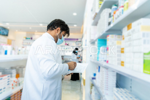 رجل صيدلاني سعودي خليجي يعمل في الصيدلية ، يرتدي الكمامة للوقاية من فايروس كورونا ،  يقوم بفحص مخزون الأدوية ويتحقق من صحتها ، الادوية والمستلزمات الطبية ، مهنة الصيدلة 