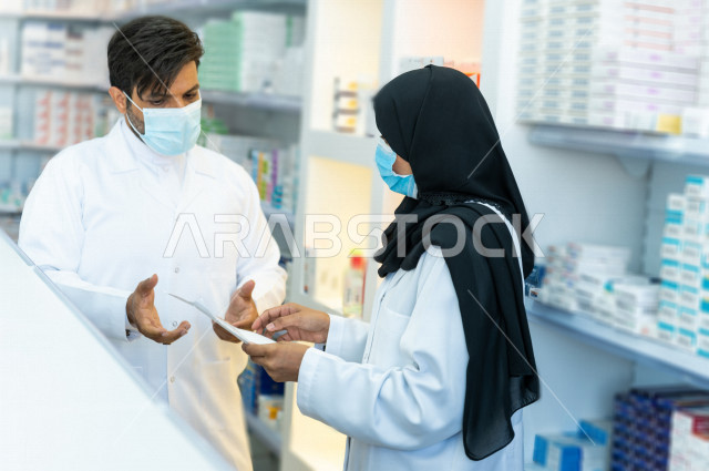 رجل وامراة عربيان خليجيان سعوديان يرتديان الكمامة للوقاية من فايروس كورونا ،  يقومان بجرد الادوية داخل الصيدلية ، مناقشة امور خاصة بالعمل ، فحص مخزون الأدوية ، تقديم الرعاية الصحية بجودة عالية ، الادوية والمستلزمات الطبية ، مهنة الصيدلة