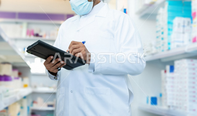 رجل صيدلاني عربي سعودي خليجي يرتدي الكمامة للوقاية من فايروس كورونا ، يقوم بجرد الادوية داخل الصيدلية ، فحص مخزون الأدوية و تدوين الملاحظات على الحافظة ، تقديم الرعاية الصحية بجودة عالية ، الادوية والمستلزمات الطبية ، مهنة الصيدلة
