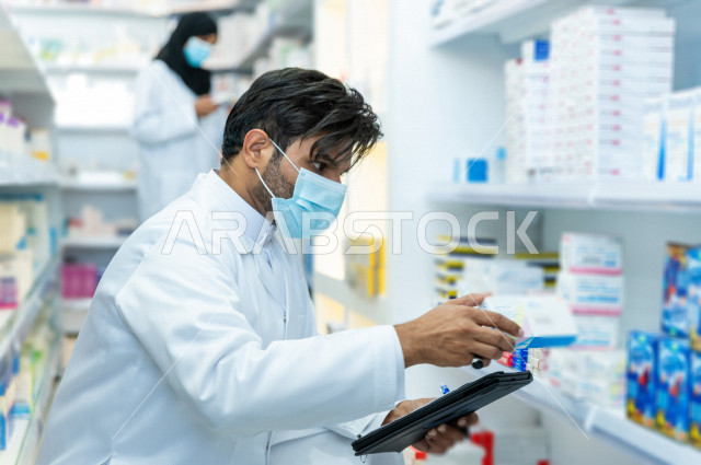 رجل صيدلاني عربي سعودي خليجي يرتدي الكمامة للوقاية من فايروس كورونا ، يقوم بجرد الادوية داخل الصيدلية ، فحص مخزون الأدوية و تدوين الملاحظات على الحافظة ، تقديم الرعاية الصحية بجودة عالية ، الادوية والمستلزمات الطبية ، مهنة الصيدلة