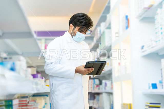 A Saudi Arabian Gulf pharmacist wearing a mask to prevent corona virus, inventorying medicines inside the pharmacy, checking the stock of medicines and taking notes on the portfolio, providing high-quality health care, medicines and medical supplies, the 
