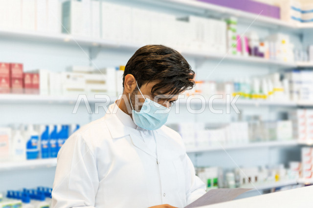 صيدلي عربي خليجي سعودي يرتدي الكمامة الطبية للوقاية من فايروس كورونا ،قراءة الروشتة الخاصة بالمريض ، صرف الادوية الموجودة في الوصفات الطبية ، توفير وتجهيز الادوية اللازمة للمريضة ، بيع الادوية والمستلزمات الطبية ، مهنة الصيدلة