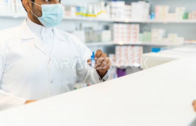 صيدلي عربي سعودي خليجي يرتدي الكمامة للوقاية من فايروس كورونا ، يحمل بيده الدواء للمريض ، صرف الادوية الموجودة في الوصفات الطبية ، توفير وتجهيز الادوية اللازمة ، بيع الادوية والمستلزمات الطبية ، مهنة الصيدلة