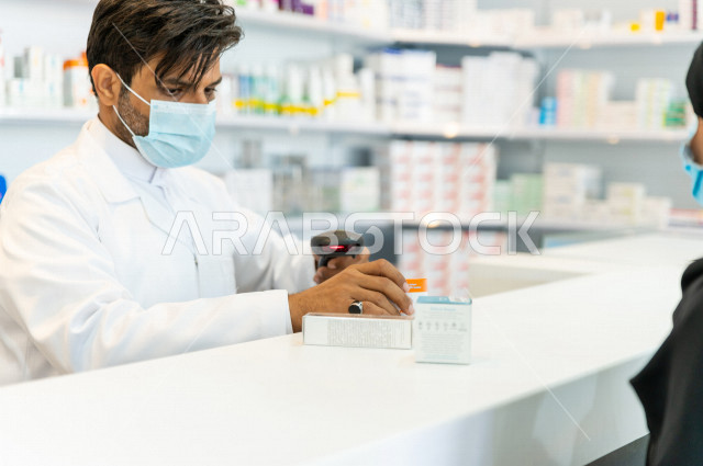A Saudi Arabian Gulf pharmacist wearing a mask to prevent corona virus, holding medicine for the patient, dispensing the medicines in the prescriptions, providing and preparing the necessary medicines, selling medicines and medical supplies, the pharmacy 