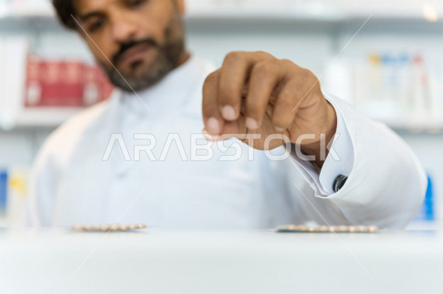 صيدلي عربي خليجي سعودي يحمل بيده قرص الدواء ،  تحضير الادوية اللازمة للمريض ، صرف الادوية الموجودة في الوصفات الطبية ، بيع الادوية والمستلزمات الطبية ، مهنة الصيدلة 