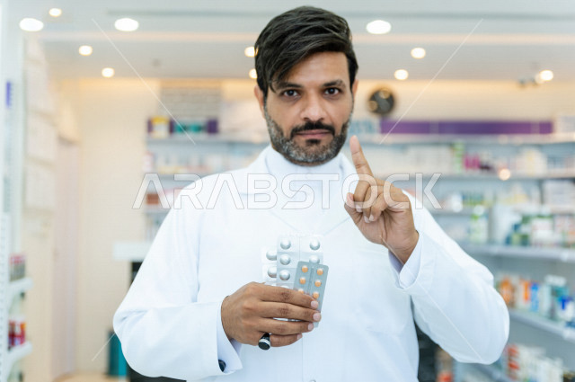 صيدلي عربي خليجي سعودي يحمل بيده اشرطة حبوب الدواء ويرفع اصبع السبابة لاعلى ، التحذير من افراط استخدام الادوية  ، صرف الادوية الموجودة في الوصفات الطبية ، بيع الادوية والمستلزمات الطبية ، مهنة الصيدلة 