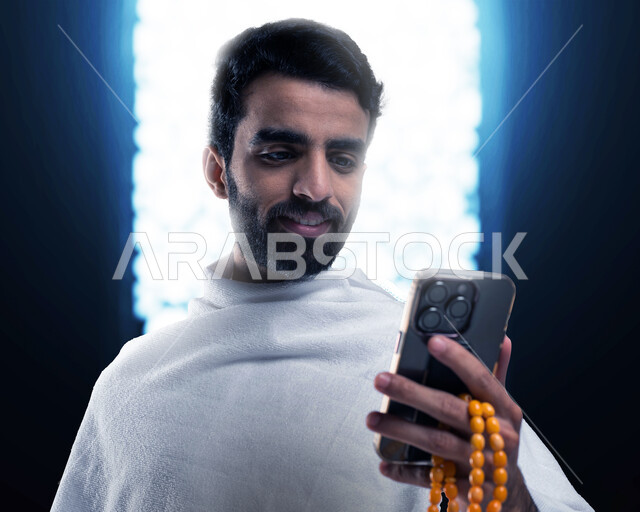استخدام تطبيق دليل الحاج والمعتمر الالكتروني على الهاتف المحمول, أداء مناسك الحج والعمرة في موسم الحج 2023, شاب سعودي خليجي مسلم يرتدي ملابس الإحرام ويستخدم الجوال ، الركن الخامس من أركان الإسلام 