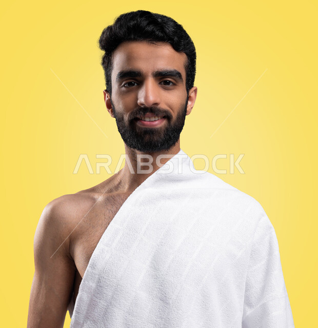 Facial gestures indicating happiness, a portrait of a Saudi Arabian Gulf Muslim young man wearing ihram clothes, looking at the camera, preparing to perform the rituals of Hajj and Umrah, the fifth pillar of Islam, Hajj season 2023, yellow background