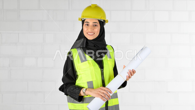 مهندسة معمارية خليجية سعودية عربية وتمسك بيدها خريطة، مخططات وتصاميم هندسية، إرتداء خوذة وسترة الحماية الخاصة بالعمل، ايماءات وجه تدل على السعادة، الاشراف على العمل