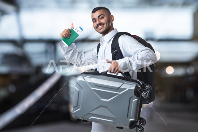 الذهاب في رحلة سياحية ممتعة، شاب سعودي خليجي يرتدي حقيبة الظهر الشخصية ويسمك بيده حقيبة السفر ويرفع اصبع الابهام لاعلى، ايماءات وجه ويدين تدل على القبول والموافقة والسعادة، الاستعداد لقضاء الاجازة الصيفية