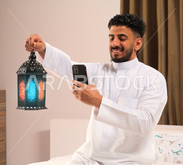 فانوس وزينة شهر رمضان، رجل خليجي سعودي عربي يمسك فانوس رمضان ويستخدم الهاتف المحمول، التقاط صور سيلفي تذكارية، فيديو بث مباشر، ايماءات وجه تدل على السعادة، اجواء رمضانية