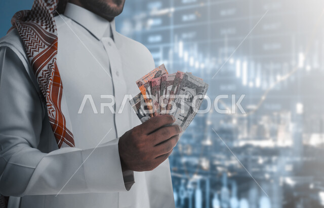 صعود وهبوط مؤشر البورصة، رجل عربي خليجي سعودي يحمل بيده كومة من العملات الورقية، إدخار وتوفير الاموال، عملات نقدية ورقية للريال السعودي، النمو والأرباح المالية، الرسم البياني للسوق المالي