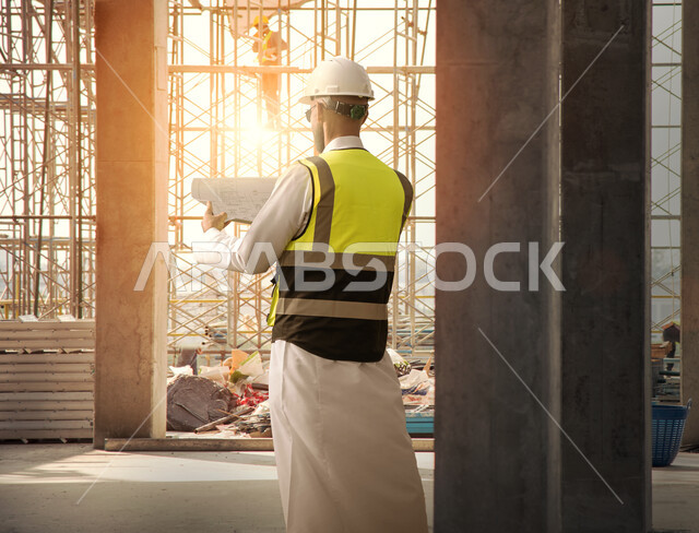 صورة من الخلف لمهندس معماري عربي سعودي خليجي يمسك بيده الخريطة ويقف في موقع البناء، مخططات وتصاميم هندسية، إرتداء خوذة وسترة الحماية الخاصة بالعمل، الاشراف على العمل