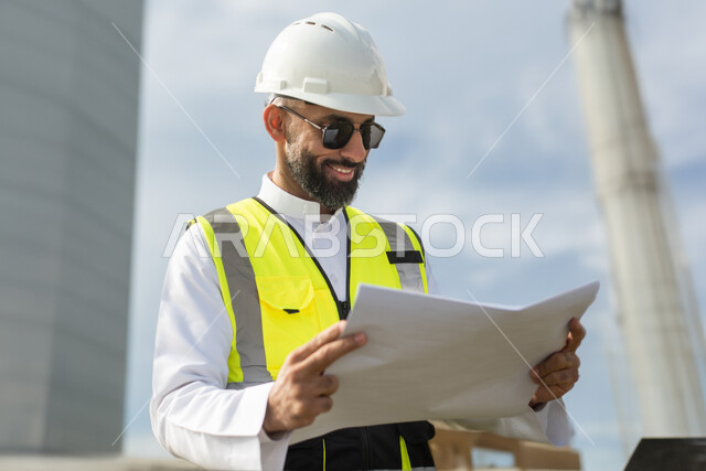 مهندس معماري خليجي سعودي عربي يرتدي نظارات شمسية سوداء ويمسك بيده خريطة، مخططات وتصاميم هندسية، إرتداء خوذة وسترة الحماية الخاصة بالعمل، ايماءات وجه تدل على السعادة، الاشراف على العمل