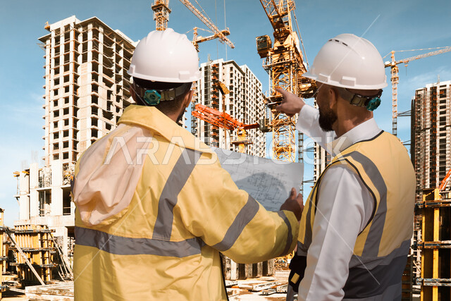 التدقيق والاشراف على العمل، مهندسان معماريان عربيان سعوديان خليجيان يتناقشان في ورقة مخطط البناء، ارتداء خوذة وسترة الحماية الخاصة بالعمل، العمل في موقع البناء، حوارات ونقاشات عمل