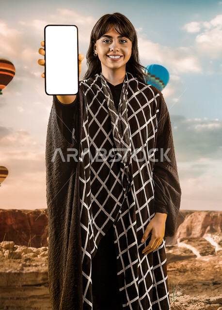 استخدام التقنية في التسويق الرقمي والالكرتوني، تطبيقات الهاتف الذكي، امرأة سعودية خليجية عربية تمسك هاتف محمول بشاشة بيضاء فارغة، اعلانات تسويقية ترويجية، بالونات الهواء الساخنة، خلفلية الطبيعة الصحراوية