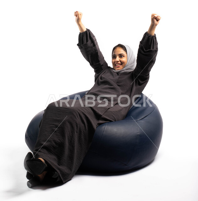 السعادة بالفوز و تحقيق الاهداف، ايماءات وجه ويدين تدل على التشجيع والحماس، بورتريه لإمرأة عربية خليجية سعودية تجلس على الكرسي ترفع يديها للأعلى وتحتفل بالفوز والانتصار، خلفية بيضاء