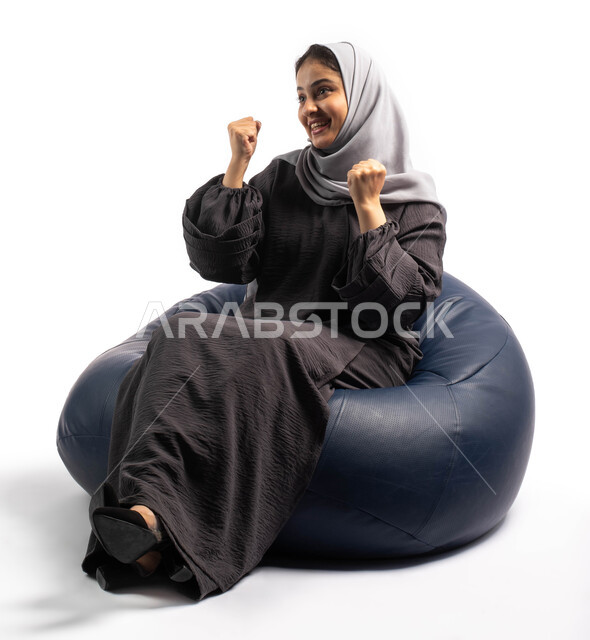 ايماءات وجه ويدين تدل على التشجيع والحماس، بورتريه لإمرأة عربية خليجية سعودية تجلس على الكرسي وتحتفل بالفوز والانتصار، السعادة بالفوز و تحقيق الاهداف، خلفية بيضاء