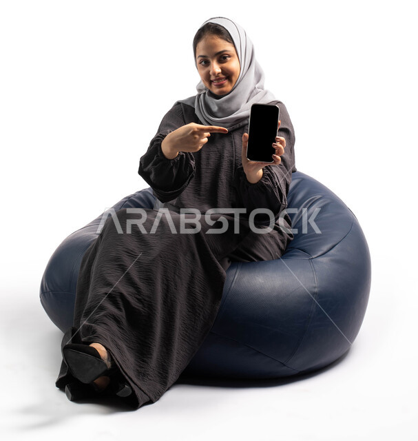 E-marketing via the Internet, displaying mobile phone services and applications, a portrait of a Saudi Arabian Gulf woman sitting on a comfortable chair and holding a mobile phone with a blank black screen and pointing at it with her finger, a white backg
