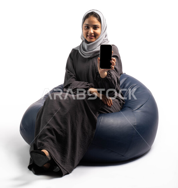 عرض خدمات وتطبيقات الهاتف الجوال، بورتريه لإمرأة عربية خليجية سعودية تجلس على كرسي مريح وتمسك بيدها الهاتف المحمول بشاشة سوداء فارغة، التسويق الإلكتروني عبر الانترنت، خلفية بيضاء