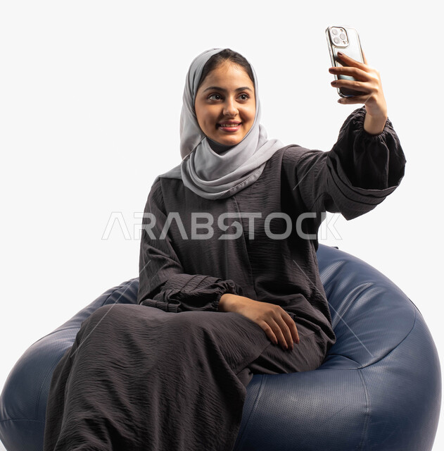 أخذ صور تذكارية على الجوال, اجراء مكالمة فيديو من خلال الهاتف الجوال، التقاط صور سيلفي, التواصل والتحدث مع الأهل والأصدقاء, بورتريه لإمرأة عربية خليجية سعودية تجلس على كرسي مريح وتمسك بيدها الهاتف المحمول، خلفية بيضاء