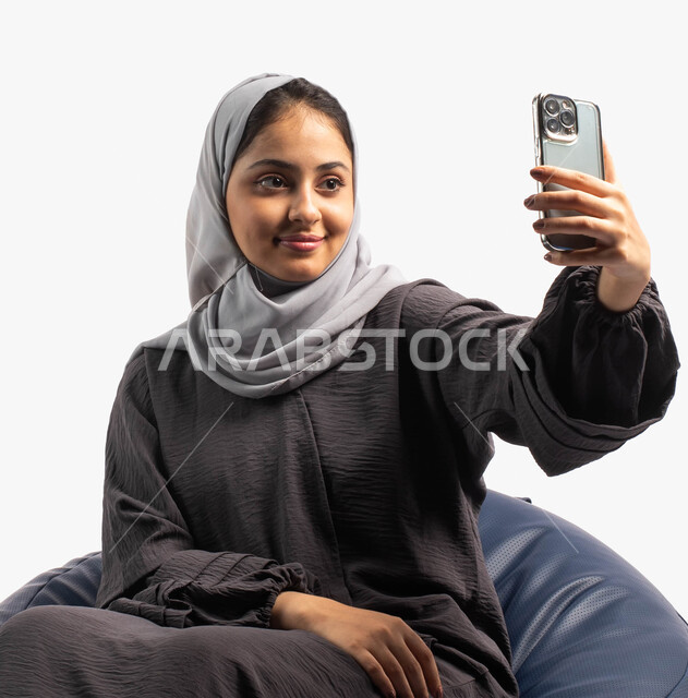 اجراء مكالمة فيديو من خلال الهاتف الجوال، التقاط صور سيلفي, التواصل والتحدث مع الأهل والأصدقاء, بورتريه لإمرأة عربية خليجية سعودية تجلس على كرسي مريح وتمسك بيدها الهاتف المحمول، خلفية بيضاء